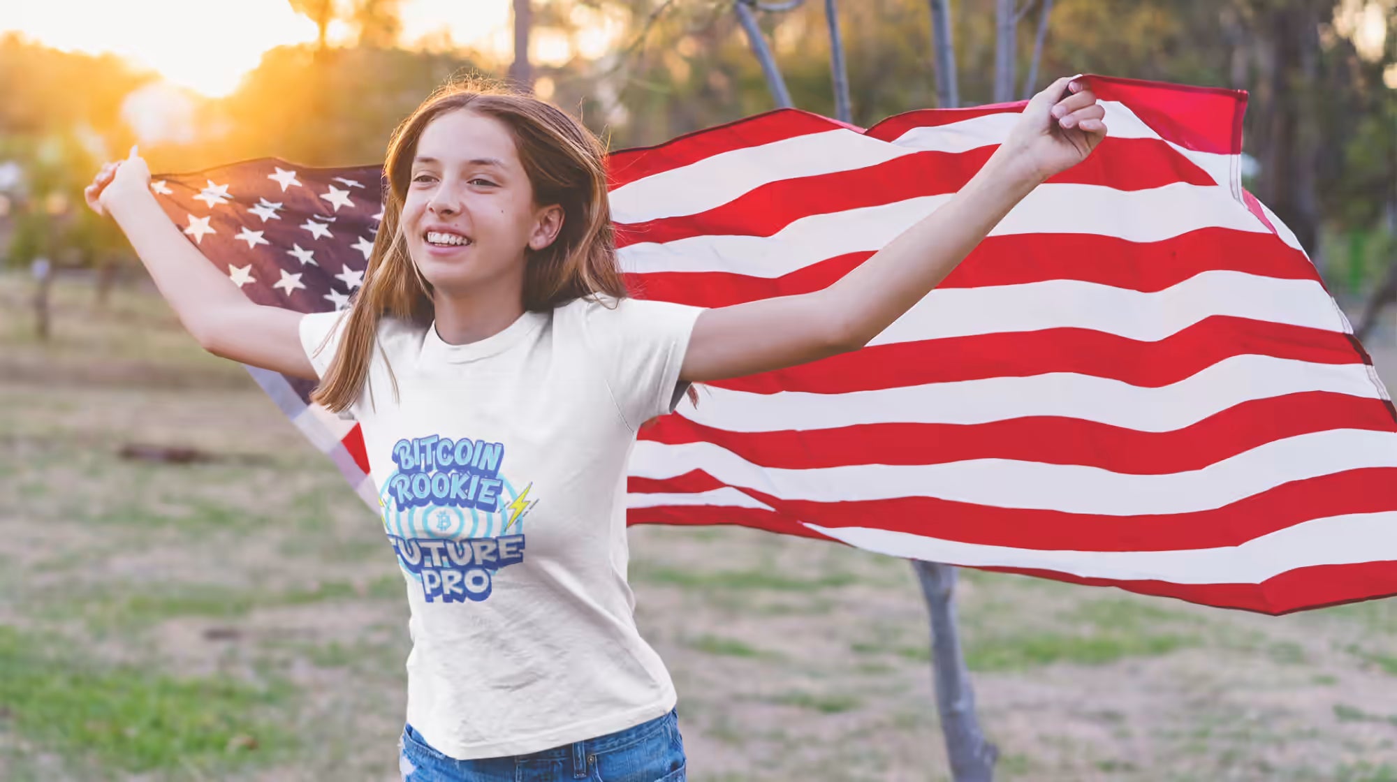 Youth T-shirts: Smiling girl in a 'Bitcoin Rookie Future Pro' t-shirt, wrapped in USA flag. Field setting.